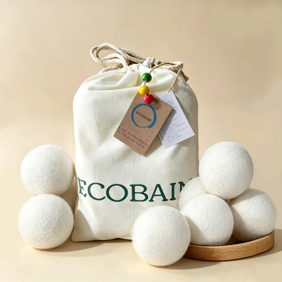 Ein Set aus sechs XXL Ecobain Trocknerbällen aus reiner, weißer Naturwolle, die dekorativ um einen hochwertigen Baumwollbeutel mit grünem Logodruck angeordnet sind. Die plastikfreie Lösung für weiche Wäsche auf einem neutralen, beigen Hintergrund