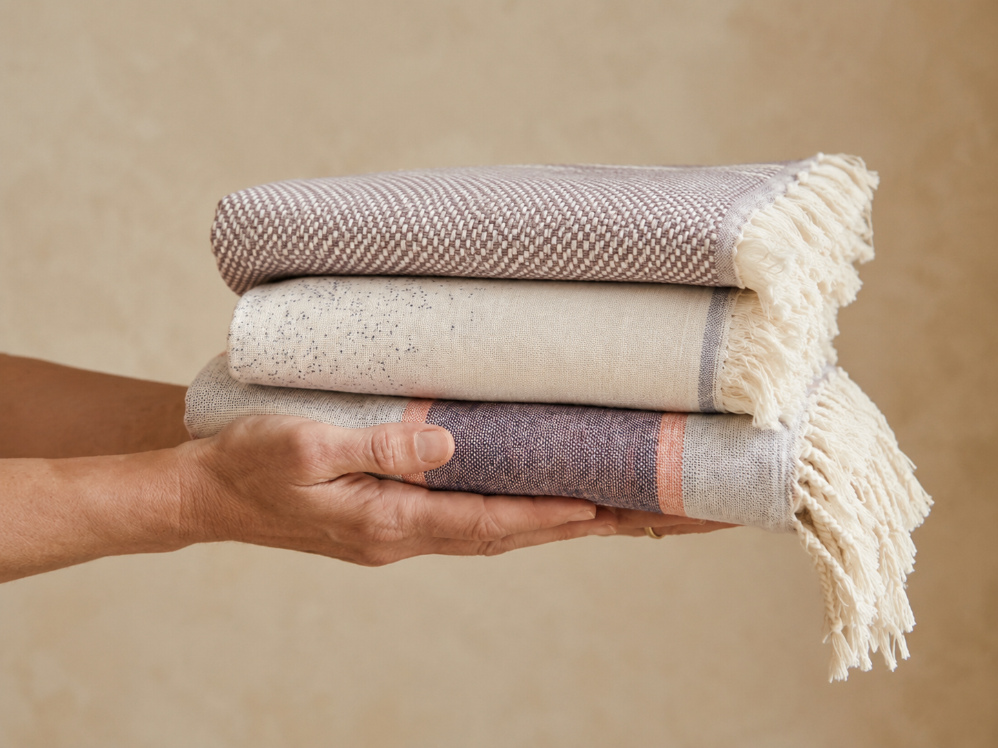 Nahaufnahme einer Hand, die einen ordentlich gefalteten Stapel aus vier Ecobain-Handtüchern präsentiert. Die Auswahl umfasst ein tealgrünes, ein fein gestreiftes naturweißes, ein terrakottafarbenes und ein senfgelbes Handtuch, alle mit der typischen, traditionell gewebten Rautenstruktur. Ein Ecobain-Papieretikett aus nachhaltigem Kraftpapier ist gut sichtbar am Stapel befestigt. Die Szene vermittelt durch den neutralen, texturierten Hintergrund und das natürliche Licht ein Gefühl von Qualität, Ordnung und p