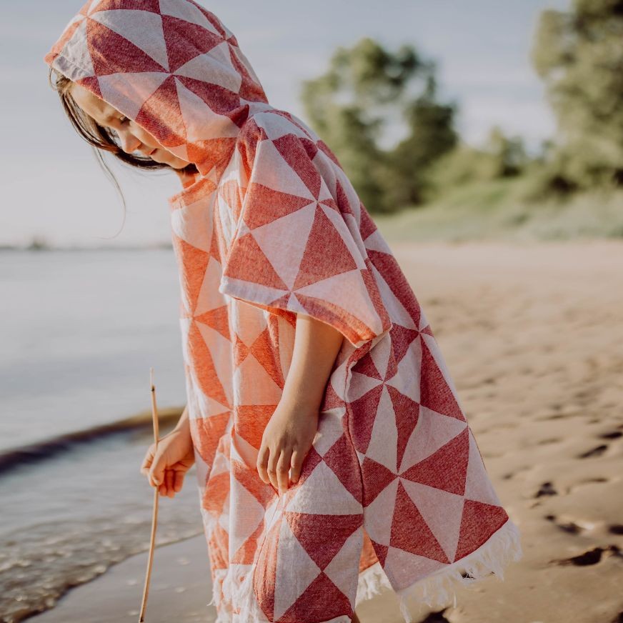 Rückansicht einer Person, die den lockeren Ecobain Poncho Symique in Wild Cherry am Strand trägt, Fokus auf den weichen Fall des Stoffes.