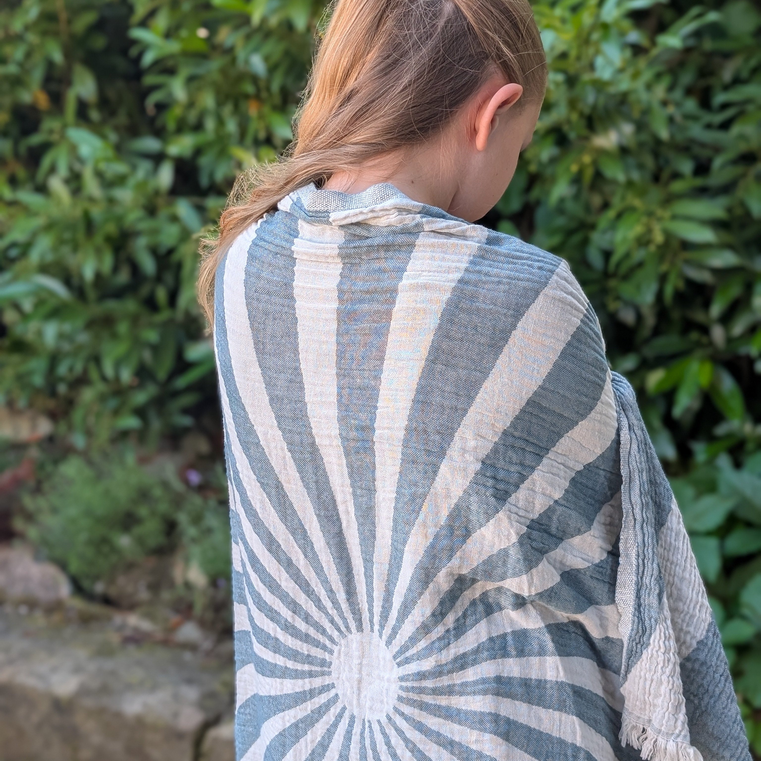 Mädchen trägt ein blau-weißes Ecobain Hammamtuch 'Sailor's Dream' mit radialem Sonnenstrahlen-Muster und feinen Fransen, lässig über die Schultern gelegt vor natürlichem Hintergrund. Hochwertige Naturfaser-Webstruktur mit weichem Fall
